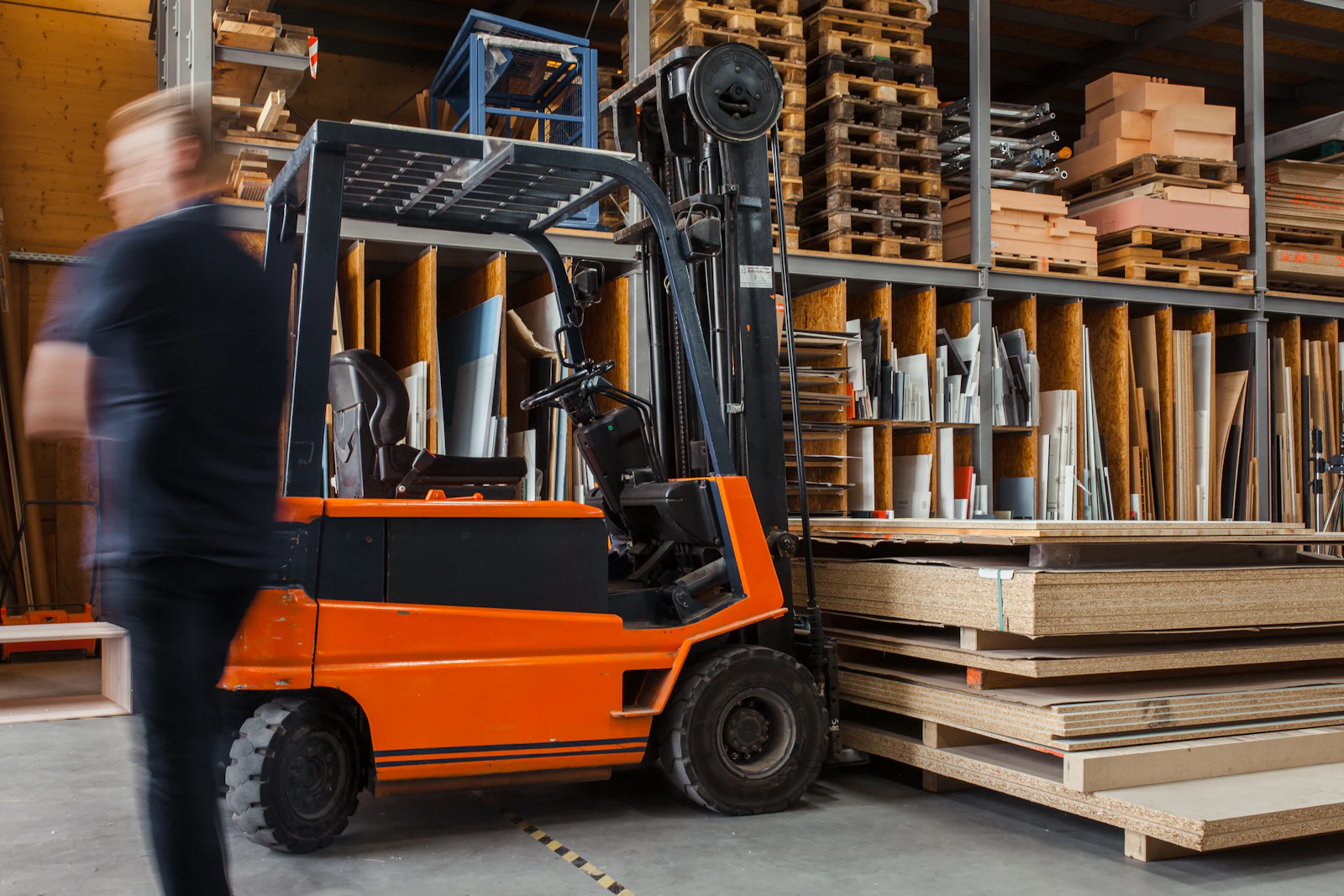 Orange forklift parked beside stacked pallets and racks of sheet goods in a warehouse