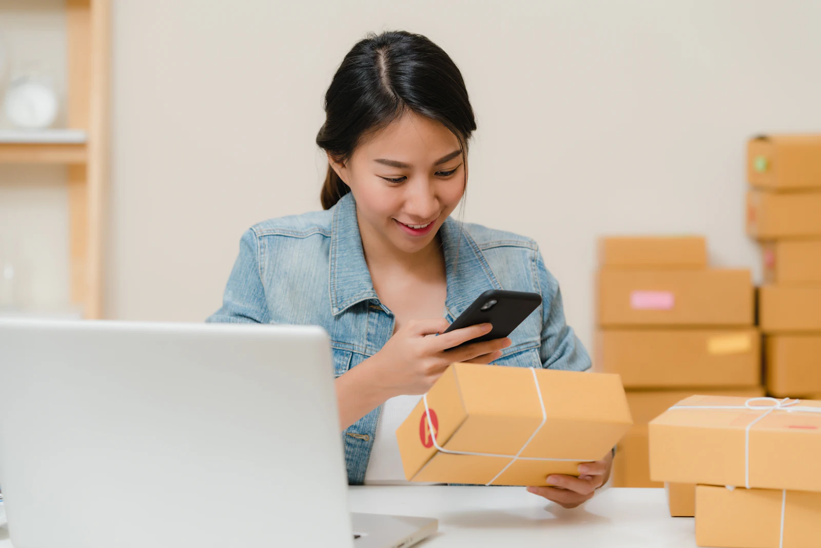 Online seller photographing a packed parcel with her phone beside a laptop and stacked boxes
